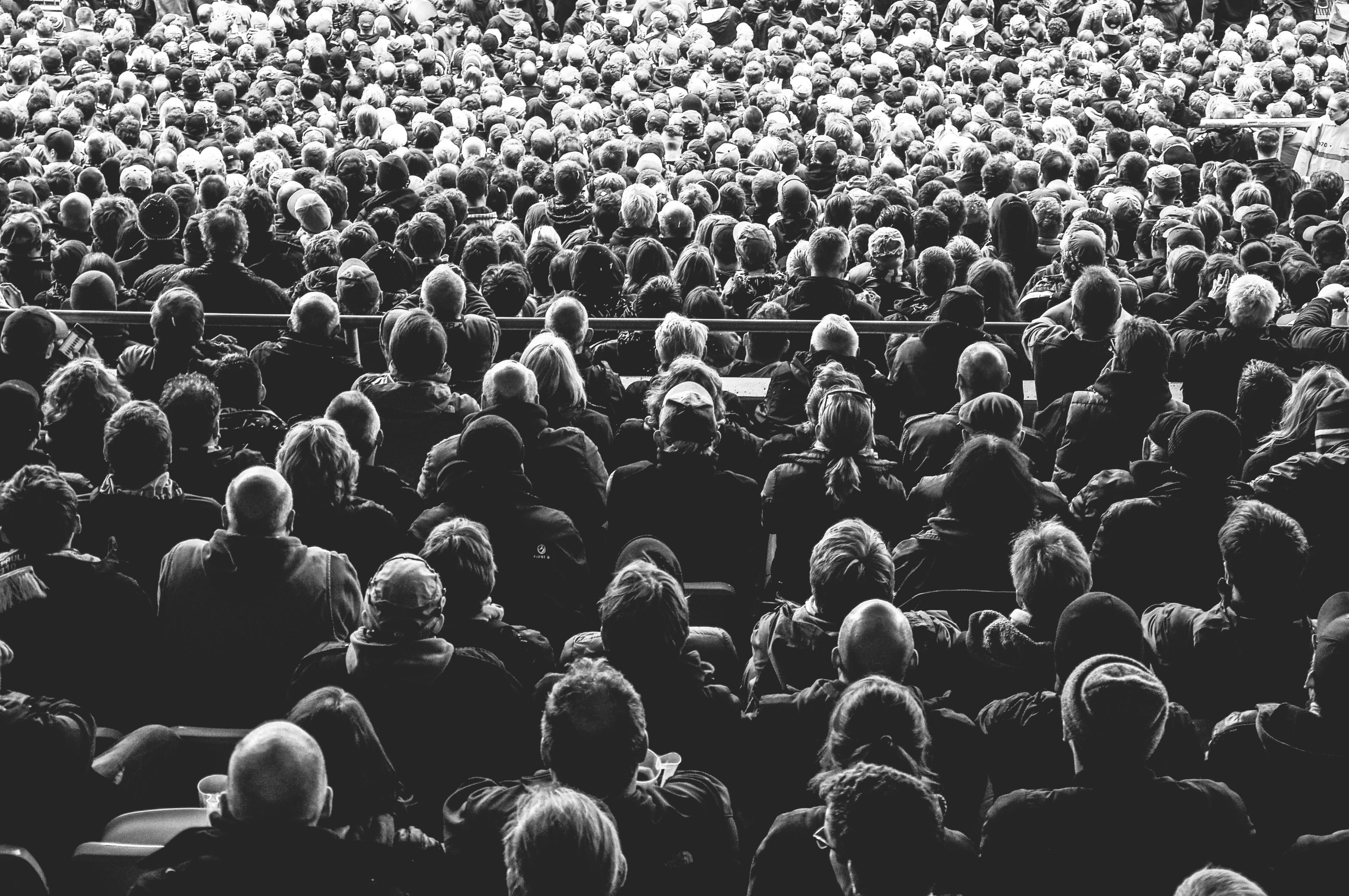 Black and white stock photo of a large audience seated and facing a brightly lit stage.